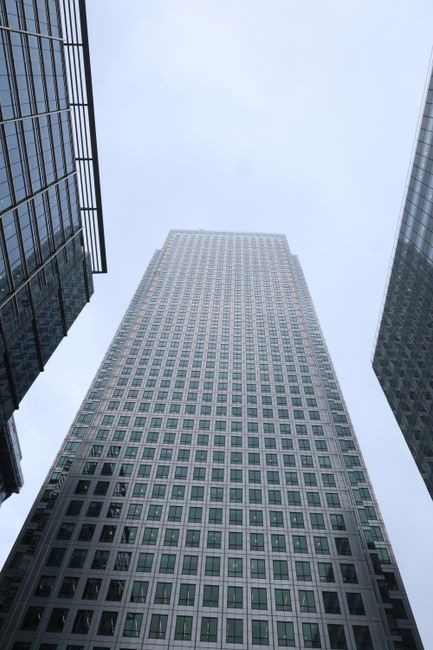 View of a tall modern skyscraper from ground level, looking directly upwards towards the grey, overcast sky. The building features a grid of windows covering its façade, with glass and metal framing. Flanking the central skyscraper are parts of two other high-rise buildings with reflective glass exteriors and dark metal structures, creating a canyon-like view typical of busy urban environments in Canary Wharf. The scene suggests a corporate or residential area, with the architecture emphasizing height and contemporary design. While the image does not include furniture or moving equipment, the setting reflects the context of house removals and furniture transport within a cityscape, fitting the theme of relocation services such as those provided by Removal Van Canary Wharf.