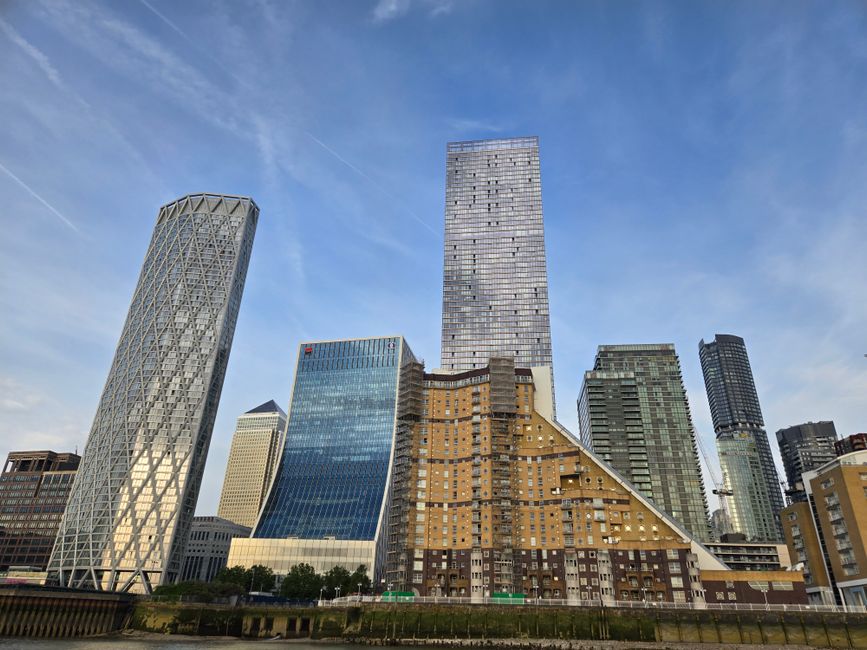A wide-angle view of the Canary Wharf skyline during daytime, featuring several modern high-rise buildings, including a prominent tall glass tower in the center, a distinctive cylindrical skyscraper on the left with a lattice exterior, and other commercial office buildings with glass facades. In the foreground, there is a construction site or building under renovation with visible construction materials such as wooden panels, scaffolding, and boarded-up sections. The area is adjacent to the River Thames, which is partially visible at the bottom of the image. The scene captures the urban landscape that home relocation services like Removal Van Canary Wharf may operate within, exemplifying the busy, high-density environment involved in packing, moving, and transporting furniture and belongings in the Canary Wharf district.