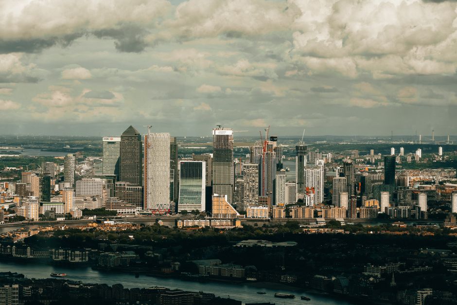 A broad view of a modern city skyline featuring numerous high-rise office buildings and residential towers under a cloudy sky. The foreground shows a dark water body with boats and low-rise structures