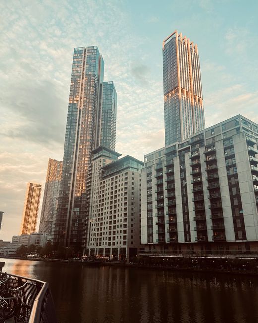 A broad view of a modern city skyline featuring numerous high-rise office buildings and residential towers under a cloudy sky. The foreground shows a dark water body with boats and low-rise structures