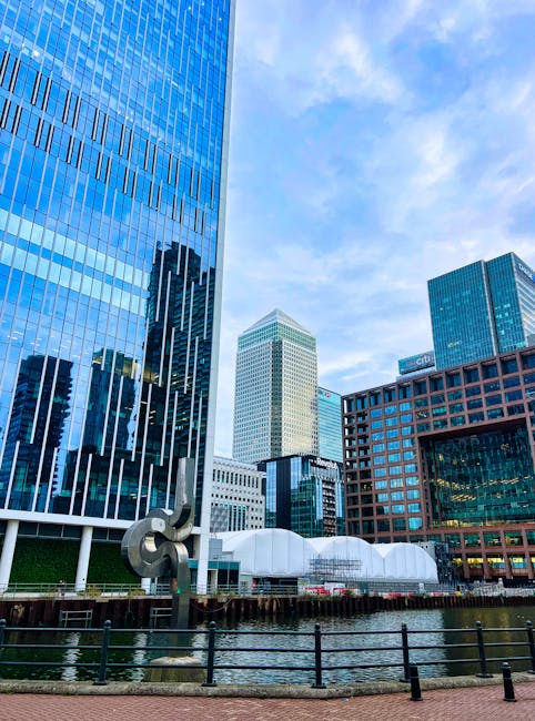 A red and white train is passing over a bridge above a river in an urban setting, with tall modern office buildings with glass facades lining the background. The scene is captured from a paved walkway