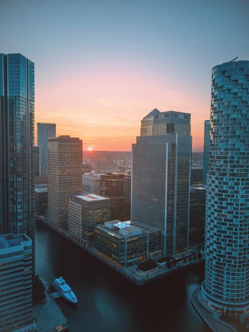 A wide aerial view of the West India Quay area in Canary Wharf during dusk, showcasing a cluster of modern high-rise office buildings and residential apartments along the riverbank. The buildings are